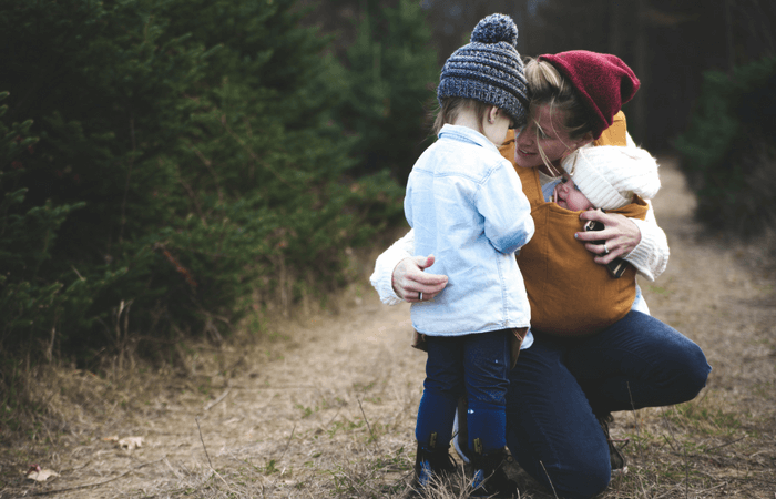 Mom holding a baby in a sling comforting a preschool child