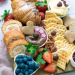 A beautifully arranged Valentine breakfast charcuterie board featuring heart-shaped waffles, pastries, bowls of cream cheese, Nutella, and small decorative toppings