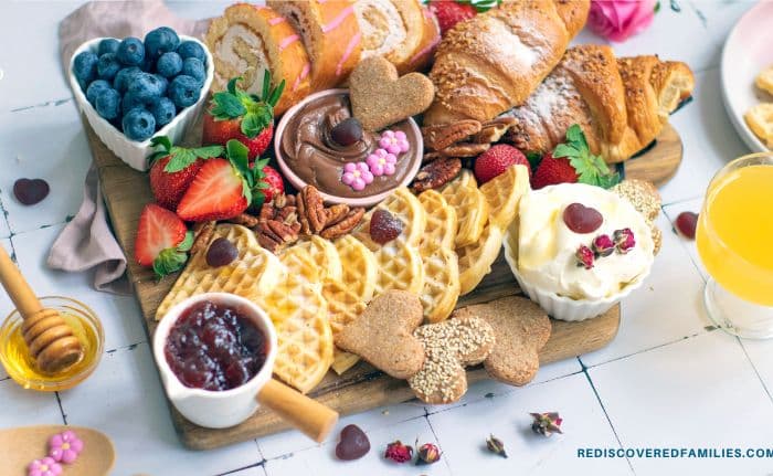 A beautifully arranged Valentine breakfast charcuterie board featuring heart-shaped waffles, pastries, bowls of cream cheese, Nutella, and small decorative toppings