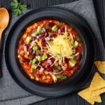 A close-up of taco soup served in a black bowl, topped with shredded cheese, avocado, and fresh herbs