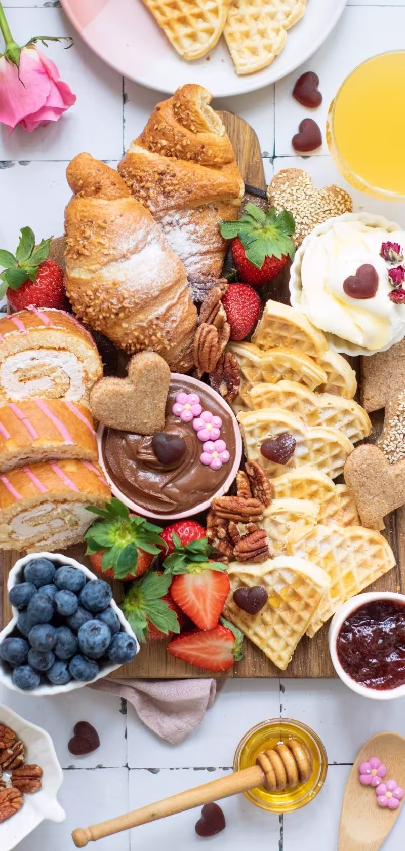 A beautifully arranged Valentine breakfast charcuterie board featuring heart-shaped waffles, pastries, bowls of cream cheese, Nutella, and small decorative toppings