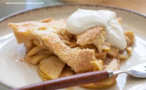 A slice of homemade apple pie with a flaky crust is served on a plate with a dollop of whipped cream.
