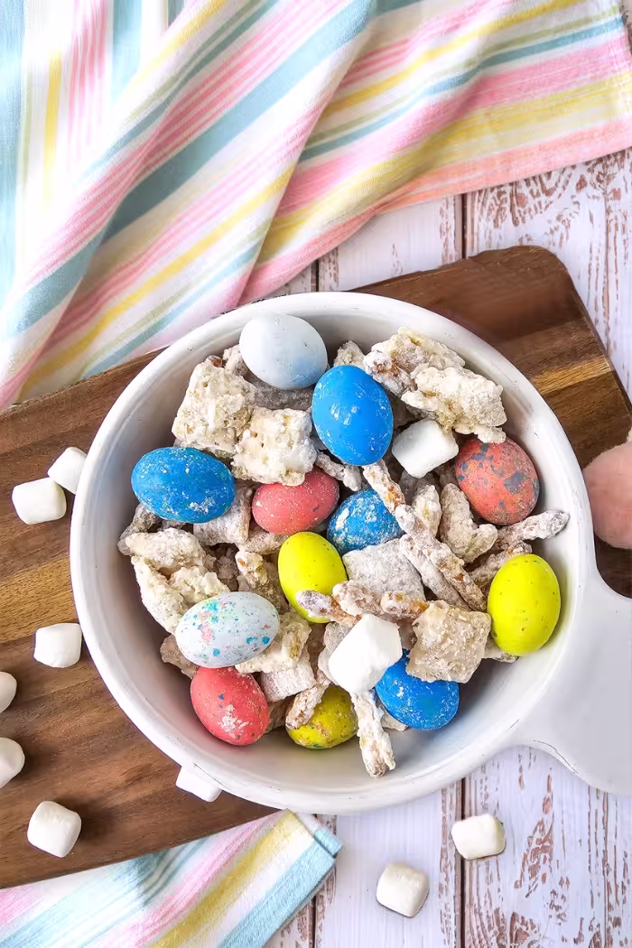 A bowl of Bunny Bait Easter snack mix placed on a wooden board with pastel-striped fabric in the background