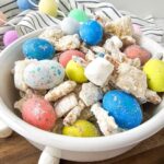 A close-up of Bunny Bait snack mix in a white bowl, with a festive striped cloth and pastel decorations in the background.