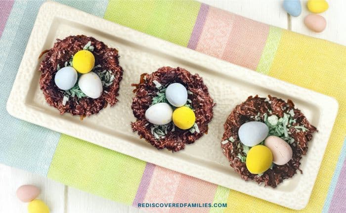 Three chocolate Easter nests with colorful candy eggs on a rectangular platter atop a pastel-striped cloth