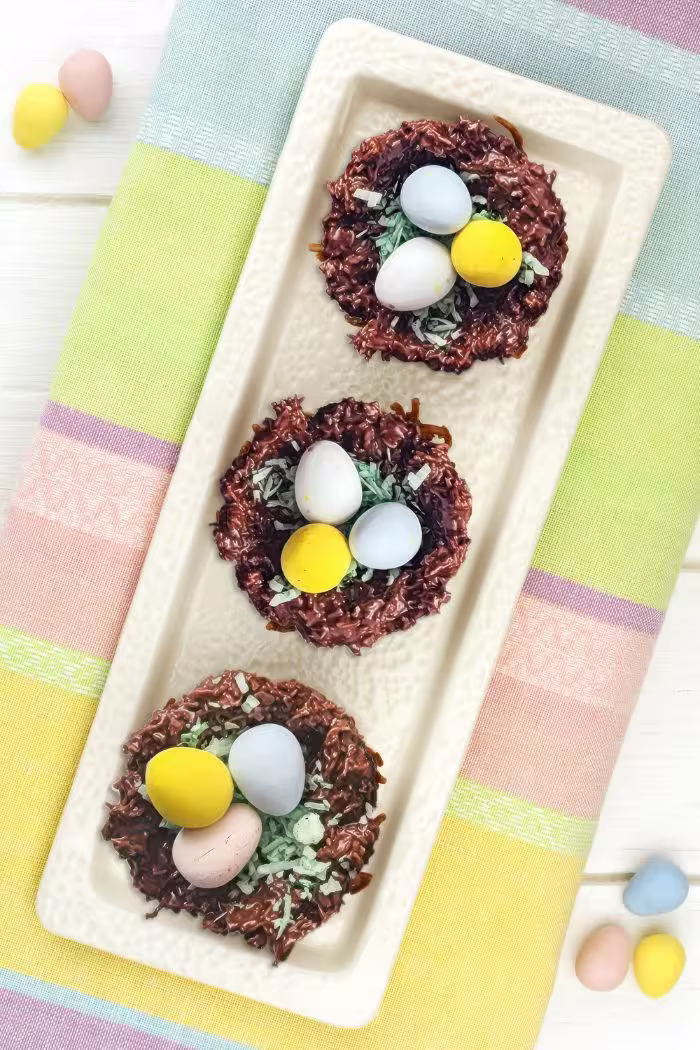 A rectangular platter holding three chocolate Easter nests filled with colorful candy eggs is displayed on a pastel-striped tablecloth.