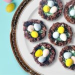A close-up of six chocolate Easter nests on a decorative plate with scattered pastel eggs