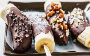 Four chocolate-coated banana halves rest on a parchment-lined tray, each topped with a different crunchy or textured ingredient, such as nuts and coconut.
