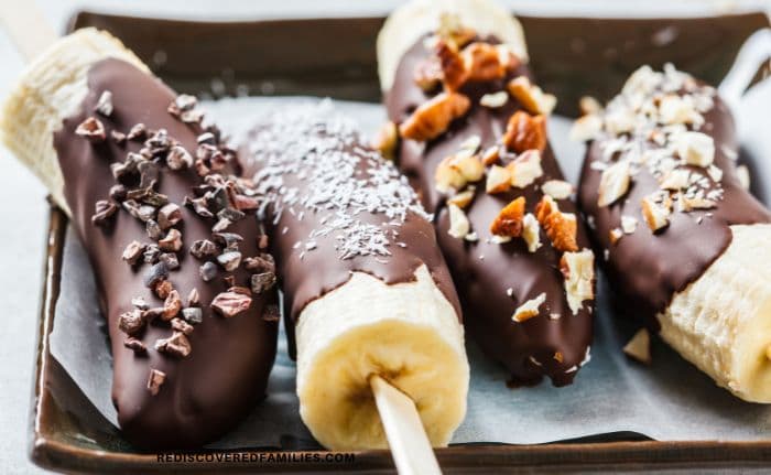 Four chocolate-coated banana halves rest on a parchment-lined tray, each topped with a different crunchy or textured ingredient, such as nuts and coconut.