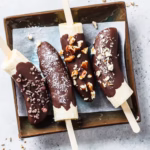Four chocolate-coated banana halves rest on a parchment-lined tray, each topped with a different crunchy or textured ingredient, such as nuts and coconut.
