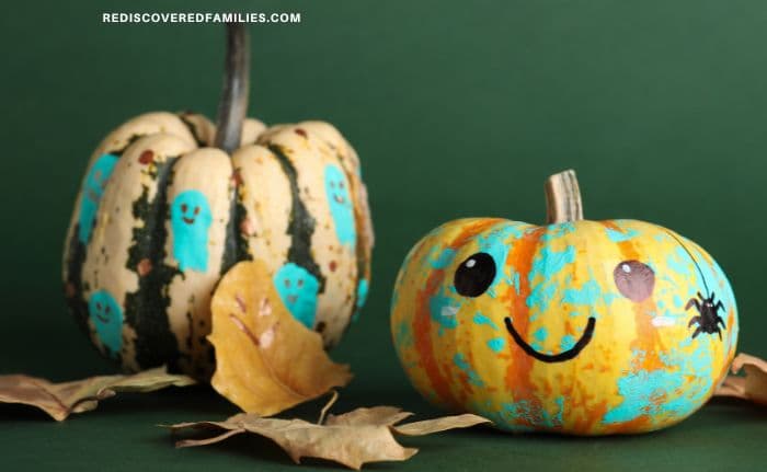 Two painted pumpkins on a green background—one with small blue ghosts on a striped gourd, the other a smiling pumpkin with splashes of turquoise, and a small painted spider.