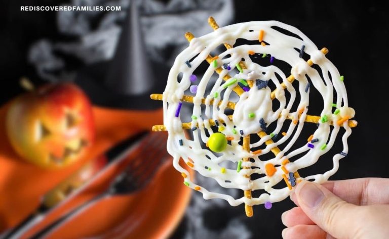 Close-up of a hand holding a Halloween chocolate pretzel spider web, with a blurred background of a jack-o'-lantern, witch hat, and orange party plates.