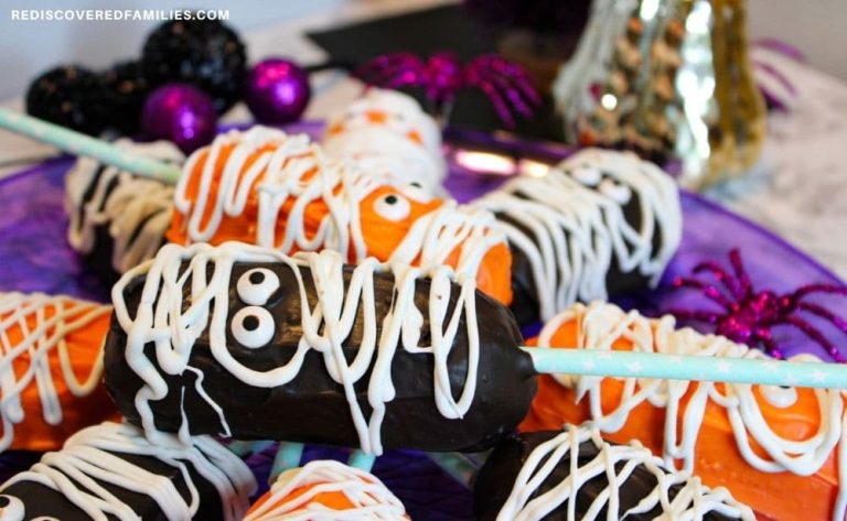 A close-up of chocolate and orange Twinkie mummies displayed on a purple tray with sparkly Halloween decorations in the background.