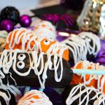 A close-up of completed Twinkie mummies in orange and chocolate, decorated with white drizzle and candy eyes, displayed on a purple tray against a backdrop of Halloween decorations.