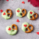 Finished Grinch pretzels scattered on a gray surface with red candy hearts.