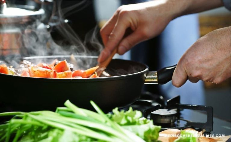 Hands stirring chopped vegetables in a steaming skillet on the stove with fresh greens in the foreground.