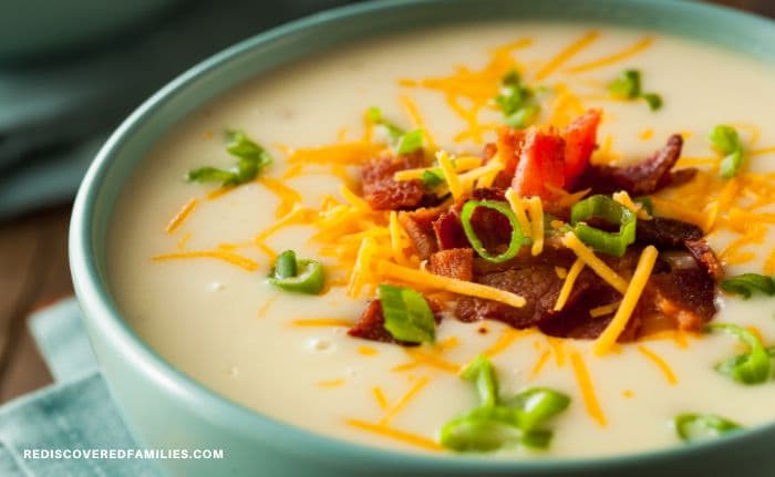 Bowl of creamy potato soup topped with bacon, cheddar cheese, and sliced green onions on a wooden table.