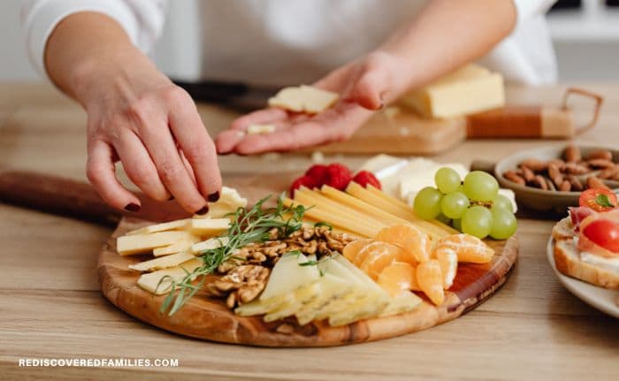Person arranging cheese, fruit, nuts, and breadsticks on a wooden board for a party snack spread.